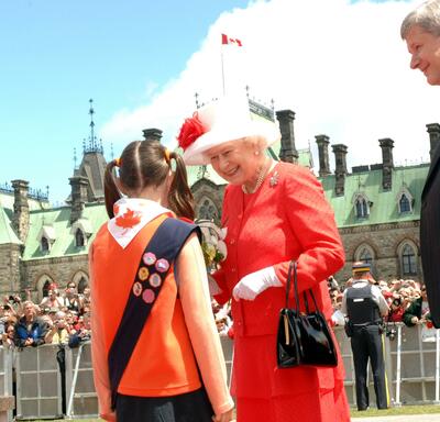 The Queen smiles as she receives flowers from a child wearing a Girl Guide uniform. Then-Prime Minister Stephen Harper looks on. A crowd of people and the Parliament Buildings are in the background.