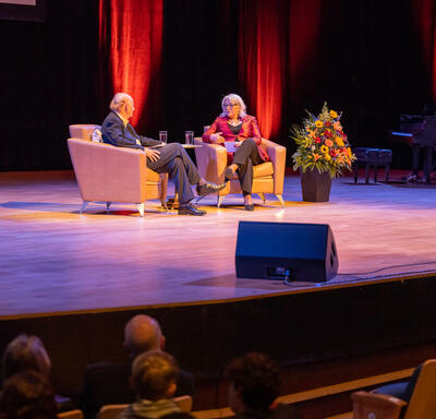 The Governor General and a man are seated in lounge chairs on a stage having a conversation.