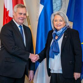 Governor General Simon shaking hands with First Deputy Speaker Antti Rinne. Behind them, in order from left to right, are the flags of Canada, Finland and the European Union.  
