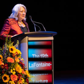 The Governor General is standing at a podium on stage. There is a lovely Fall floral arrangement to her left.