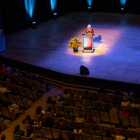 Vue du balcon de la salle de concert Jack Singer à Calgary. La gouverneure générale est debout à un pupitre sur la scène.