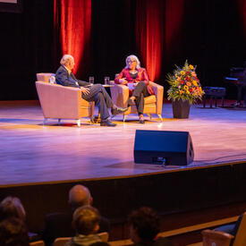 The Governor General and a man are seated in lounge chairs on a stage having a conversation.