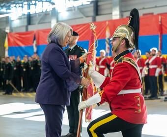 Governor General Mary Simon wears a purple suit and smiles at a male member of the Canadian Armed Forces as she presents the guidon. The member of the Canadian Armed Forces wears a red uniform, white gloves, and a helmet.