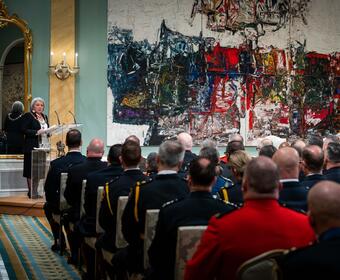 Governor General Mary Simon speaks at a podium in the ballroom at Rideau Hall. She is looking out at a crowd of recipients of the Order of Merit of the Police Forces. The recipients are wearing police uniforms. 